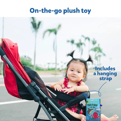 A baby in a red stroller holding a soft book with a hanging strap.