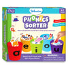Front view of the Skillmatics Phonics Sorter game box for ages 4 to 7, featuring five large cups labeled a, e, i, o, and u filled with illustrated picture pieces like cat, bed, pig, and cup. Text explains phonetic sound sorting and mastering five vowels with over 100 picture pieces. The design highlights speech, listening skills, phonemic awareness, and hands-on early learning play.