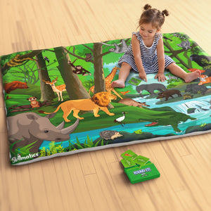 A toddler sits barefoot on a large, illustrated jungle-themed cloth play mat placed on a wooden floor, pointing toward animal illustrations. The mat features a detailed forest scene with a lion, elephant, giraffe, crocodile, rhinoceros, deer, tiger, birds, monkey, sloth, and flowing river. A small green game card and an open Skillmatics Found It box lie nearby.