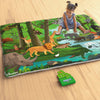 A toddler sits barefoot on a large, illustrated jungle-themed cloth play mat placed on a wooden floor, pointing toward animal illustrations. The mat features a detailed forest scene with a lion, elephant, giraffe, crocodile, rhinoceros, deer, tiger, birds, monkey, sloth, and flowing river. A small green game card and an open Skillmatics Found It box lie nearby.