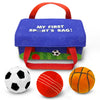 Blue fabric children’s sports bag with red carry handles displayed on a white background. The bag reads “My First Sport’s Bag!” and features subtle sports ball patterns and illustrated grass along the bottom edge. In front of the bag are three plush balls: a black-and-white soccer ball, a red cricket ball with stitched lines, and an orange basketball with black detailing.