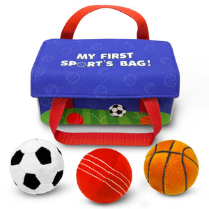 Blue fabric children’s sports bag with red carry handles displayed on a white background. The bag reads “My First Sport’s Bag!” and features subtle sports ball patterns and illustrated grass along the bottom edge. In front of the bag are three plush balls: a black-and-white soccer ball, a red cricket ball with stitched lines, and an orange basketball with black detailing.