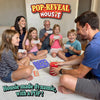 A family of four sits at a table playing Pop-Reveal Housie. An adult man holds the red pop-reveal can while smiling, with a woman and two children holding housie cards and watching. A blue number tray with yellow balls sits on the table. The Pop-Reveal Housie logo appears above, with text reading “Housie made dynamic, with a POP!”