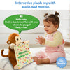 A smiling baby in pastel pink overalls and a yellow striped top sits on the floor, clapping while facing a plush monkey toy holding a polka-dotted cloth printed with “Peek-a-boo, where are you?” A green speech bubble shows the toy’s audio phrase. Blue text above reads “Interactive plush toy with audio and motion,” and a “12+ months” badge appears in the corner.