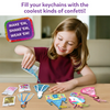 A smiling child sits at a table pouring blue confetti from a small packet into a unicorn-shaped foam shake-it. On the tabletop are additional foam shapes, transparent sheets, keychain loops, and multiple confetti packs in pink, gold, and multicolor. A purple badge reads “Make ’em, shake ’em, wear ’em,” while text above says “Fill your keychains with the coolest kinds of confetti.”