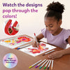 A young girl seated at a wooden table smiling while coloring Paint-Over Art sheets with markers; completed bright floral and fantasy designs visible, open kit box and scattered markers around, text reads Watch the designs pop through the colors, Magic Shine-Through badge shown.