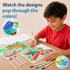 Child sitting at table smiling while using paint-over art markers on illustrated activity sheets, revealing bright colors through hidden patterns; multiple markers laid out on wooden surface and finished colorful animal designs visible beside ongoing coloring activity.