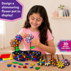 Lifestyle image of a child creating Skillmatics Foil Fun Mosaics 3D flower pot craft. The child attaches glittery foil flower pieces while a completed colourful flower pot with sparkling flowers and leaves sits on the table. Includes text highlighting a shimmering flower pot display and 3D mosaic foil craft.