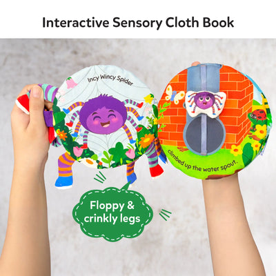 Hands holding an open Incy Wincy Spider interactive sensory cloth book. The left page shows a cheerful purple spider illustration with colorful legs and flowers, while the right page features a brick wall scene with a fabric spider that moves along a vertical strap. A callout highlights floppy and crinkly legs. The soft fabric pages, stitched details, and movable elements encourage tactile play and exploration.