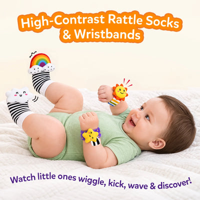 Baby lying on a soft white bed wearing Skillmatics Wriggle & Wonder high-contrast socks and wristbands, smiling while kicking and moving. Rainbow, cloud, sun and star designs support visual and sensory discovery, with the heading “High-Contrast Rattle Socks & Wristbands.”
