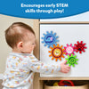 A toddler in patterned pajamas plays with the Skillmatics Spinny Gears toy on a cabinet surface. The colourful gears interlock and spin, each featuring a different animal. The caption reads “Encourages early STEM skills through play!” highlighting how the toy promotes cause-and-effect learning, hand-eye coordination, and sensory exploration.