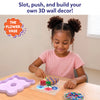 Young child seated at a table assembling the Skillmatics Stixels Flower Vase craft by inserting colorful Stixels into a transparent flower-shaped base placed over a color guide. A lavender frame sits nearby, with extra Stixels organized in a bowl beside the partially completed 3D design.