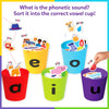 Layout showing contents of the Skillmatics Phonics Sorter game, including five plastic vowel cups labeled a, e, i, o, and u, over 100 illustrated picture pieces featuring animals, people, objects, and everyday items, and a printed learning guide. The image clearly displays each component separately on a clean background, helping parents understand what is included in the phonics learning kit.