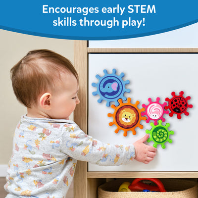 A toddler in a grey floral top reaches to touch Skillmatics Spinny Gears mounted on a white cabinet door. Five interlocking animal gears are visible: blue elephant, orange lion, pink rabbit, red ladybug, and green caterpillar. A toy basket sits below the cabinet. Header reads "Encourages early STEM skills through play!"