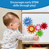 A toddler in a grey floral top reaches to touch Skillmatics Spinny Gears mounted on a white cabinet door. Five interlocking animal gears are visible: blue elephant, orange lion, pink rabbit, red ladybug, and green caterpillar. A toy basket sits below the cabinet. Header reads 