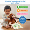 A smiling baby in printed pajamas sits on the floor gently pressing the tail of a brown plush monkey toy. The monkey holds a polka-dotted cloth printed with “Peek-a-boo, where are you?” At the top, blue text reads “Press the monkey’s tail to play peek-a-boo!” Icons on the right show “Battery Operated,” “Soft & Fuzzy,” and “Do Not Wash” in green, orange, and pink labels.