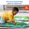 A young child wearing a yellow shirt and blue pants kneels on a jungle-themed cloth play mat, holding a green animal card and placing it over a matching illustration on the mat. The mat shows a river, crocodile, elephant, birds, and forest animals. Text above reads “First ever search & find game on a play mat!” with an “18+ months” age badge.
