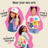 Three children showcasing Skillmatics Iron-Its wearable art. Left: girl wearing white bucket hat with cherry iron-on design. Right: girl in white tee featuring large colorful flower print. Center-bottom: girl carrying canvas tote bag decorated with colorful seashell and ocean designs.