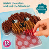 Close-up instructional view of Skillmatics Stixels 3D art activity showing a hand placing a pink stixel peg into a transparent base board to form a puppy face design. The image emphasizes color matching, slot-and-fill technique, with cylindrical pegs creating a textured mosaic effect.