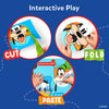 A blue background graphic titled “Interactive Play” showing three circular steps. The first shows a hand using green scissors to cut around a Disney character face. The second shows hands folding a cut paper shape. The third shows a child applying glue with a Skillmatics glue stick to paste pieces together. Labels reading “Cut,” “Fold,” and “Paste” guide the activity flow visually.