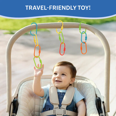 Baby seated in a padded stroller reaches upward to touch colorful plastic Linkies hanging from the stroller handle. The links in yellow, green, blue, orange, and red are clipped individually and in short chains. A blue banner at the top reads “Travel-Friendly Toy.” The background shows a softly blurred outdoor path, emphasizing on-the-go play and reach-and-grasp interaction.