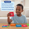 A young child sits at a table holding a red “This or That” Skillmatics spelling card while smiling and looking at it. Multiple stacks of colorful flashcards labeled Tricky Spellings, General Rules, Mnemonics, and This or That are neatly placed on the table. A blue text panel lists learning outcomes such as spelling pattern recognition, letter-sound associations, word recall, and vocabulary development.