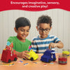 Two children sit at a table shaping Sandeezy kinetic-sand vehicles, decorating a red fire-rescue truck, a yellow taxi, and a blue ice-cream van using colorful signs, cones, lights, and luggage pieces spread across the table. Jars of red and yellow sand are placed near them. The top banner reads “Encourages imaginative, sensory, and creative play!” on a curved red background.