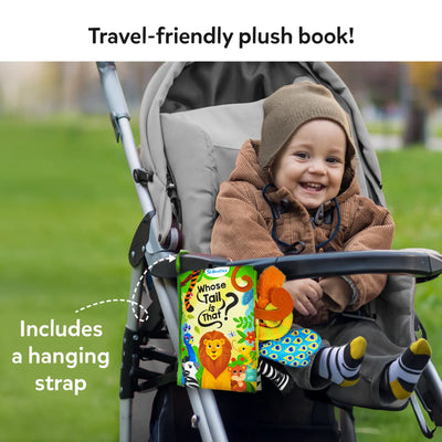 A baby seated in a stroller, wearing a cozy winter jacket, a knitted hat, and striped socks, smiling joyfully. Attached to the stroller is the 'Whose Tail is That?' cloth book, featuring vibrant illustrations of animals like a lion, zebra, and peacock, with colorful plush tails hanging from the book. The book has a convenient hanging strap, making it travel-friendly for parents and babies.
