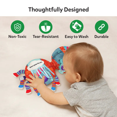 A baby lying on their stomach reaches out to touch the Incy Wincy Spider sensory cloth book placed on a soft surface. The book is open, showing colorful illustrated pages with a water spout scene and fabric spider elements. Above the baby, icons and text highlight non-toxic materials, tear-resistant construction, easy-to-wash fabric, and durable design, visually reinforcing safe and thoughtful product features.