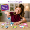 A smiling child sits at a table pouring blue confetti from a small packet into a unicorn-shaped foam shake-it. On the tabletop are additional foam shapes, transparent sheets, keychain loops, and multiple confetti packs in pink, gold, and multicolor. A purple badge reads “Make ’em, shake ’em, wear ’em,” while text above says “Fill your keychains with the coolest kinds of confetti.”