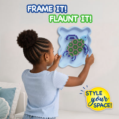 Child placing a completed Skillmatics Stixels Sea Turtle artwork onto a bedroom wall. The framed 3D wall decor features colorful Stixels arranged on a turtle-shaped design inside a light blue frame. The image showcases the finished DIY craft as personalized room décor for kids.