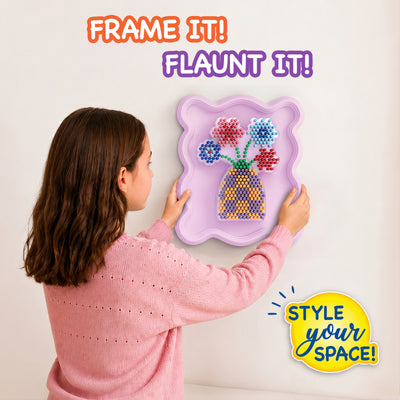 Child wearing a pink sweater hangs a completed Skillmatics Stixels Flower Vase artwork on a wall. The finished 3D design features colorful flowers and a patterned vase mounted inside a lavender decorative frame, demonstrating how the craft transforms into personalized wall décor for a bedroom or playroom.