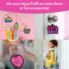 Lifestyle collage showing finished Aqua Puffs used as décor and accessories. Designs hang on pink wire wall grid beside pens and clock, attached to door knob with yellow string, held as keychain in hand, and clipped to yellow backpack worn by smiling child. Text reads: Use your Aqua Puffs as room decor or fun accessories.