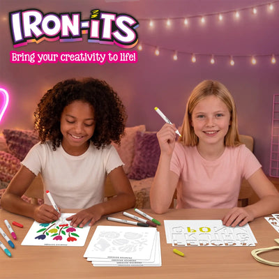 Two girls craft together at a wooden table in a pink room with fairy lights. Left girl with curly hair colors a floral stencil with a red iron-on marker. Right girl holds a yellow marker and smiles. Iron-Its logo reads: Bring your creativity to life!