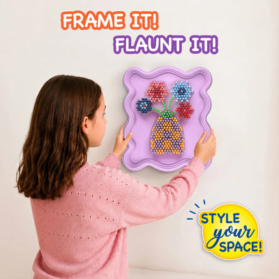 Child wearing a pink sweater hangs a completed Skillmatics Stixels Flower Vase artwork on a wall. The finished 3D design features colorful flowers and a patterned vase mounted inside a lavender decorative frame, demonstrating how the craft transforms into personalized wall décor for a bedroom or playroom.