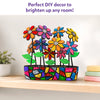Completed DIY mosaic flower pot displayed on a wooden shelf, featuring colorful glittery flowers, bright green leaves, and a vibrant mosaic-patterned planter. Soft home décor, books, and star wall accents in the background highlight the craft as a decorative display piece for any room.