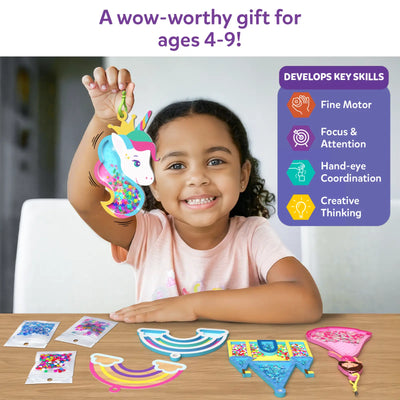 A smiling child sits at a table holding a unicorn shake-it keychain filled with multicolored confetti. On the table are rainbow and castle foam shake-its, confetti packs, and keychain parts. A side panel titled “Develops Key Skills” lists icons for fine motor skills, focus and attention, hand-eye coordination, and creative thinking, with text stating it is suitable for ages 4–9.