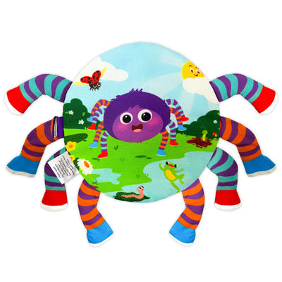 A circular fabric page featuring a smiling purple spider illustration set in a bright outdoor scene with grass, puddles, flowers, and small animals like a frog and worm. The spider has eight soft, striped fabric legs extending outward in multiple colors. A stitched label is visible on the side. The flat, padded page and protruding legs emphasize tactile exploration and visual storytelling through fabric textures.