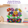 Completed DIY mosaic flower pot displayed on a wooden shelf, featuring colorful glittery flowers, bright green leaves, and a vibrant mosaic-patterned planter. Soft home décor, books, and star wall accents in the background highlight the craft as a decorative display piece for any room.