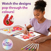 A young girl seated at a wooden table smiling while coloring Paint-Over Art sheets with markers; completed bright floral and fantasy designs visible, open kit box and scattered markers around, text reads Watch the designs pop through the colors, Magic Shine-Through badge shown.